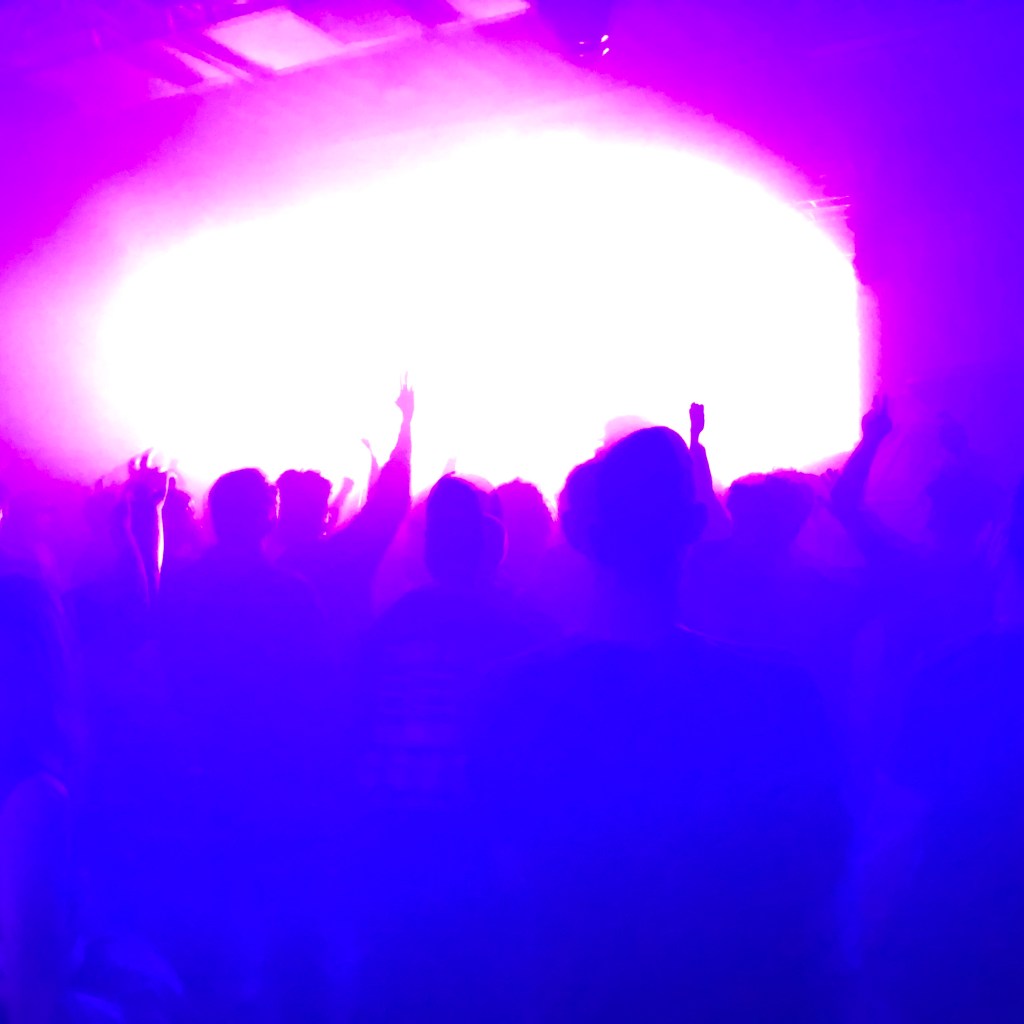 Light flare inside a rave silhouettes arms in the air of people dancing.